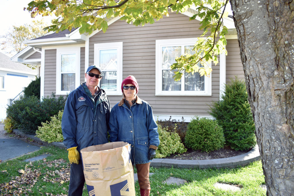 A couple standing in their front yard with a large bag of leaves on a Fall day in St. John's.