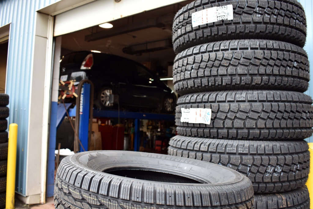 A pile of tires is in the forefront of an auto service station. In the background a car is up on a ramp having its tires installed for winter.
