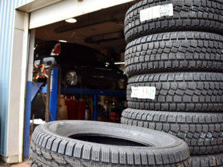 A pile of tires is in the forefront of an auto service station. In the background a car is up on a ramp having its tires installed for winter.