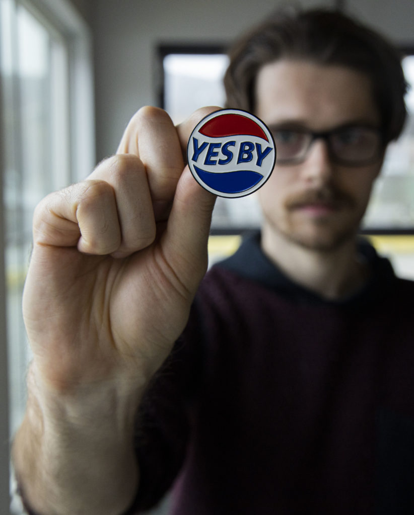 Etsy shop owner Alex Wilkie holds up an enamel pin that says "Yes By."