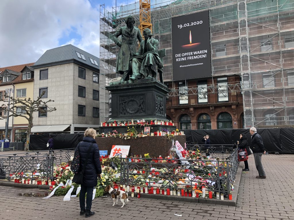 People stand around an impromptu memorial for the victims of the Hanau shooting. Wreaths and candles have been laid down around the sculpture of Brothers Grimm on the marketplace. A banner on town hall reads "The victims weren't strangers. #hanaustandstogether". Photo courtesy of Hessischer Rundfunk
