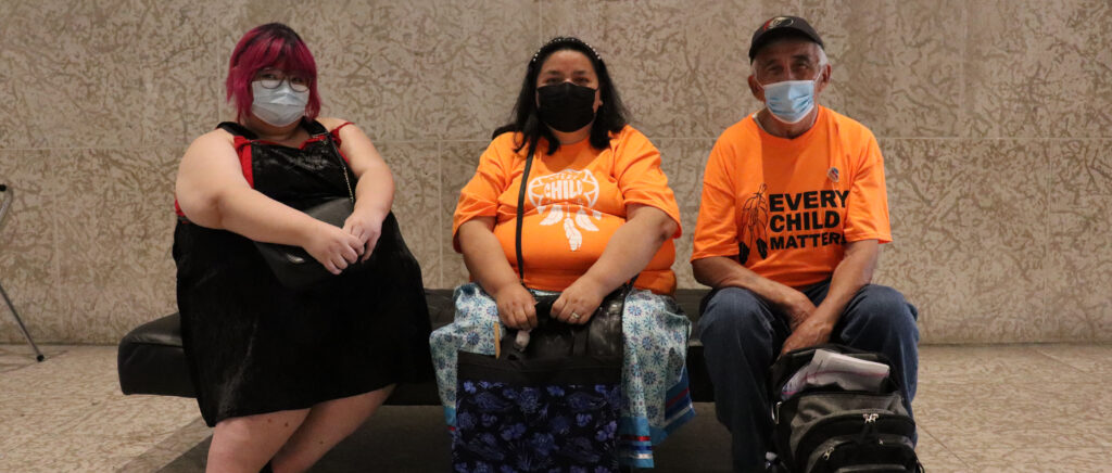 Three masked adults, two wearing orange shirts sit bench gallery