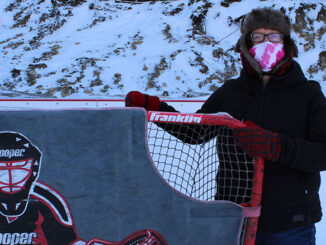 woman in winter clothes stands by hockey net on outdoor rink with snow in the background