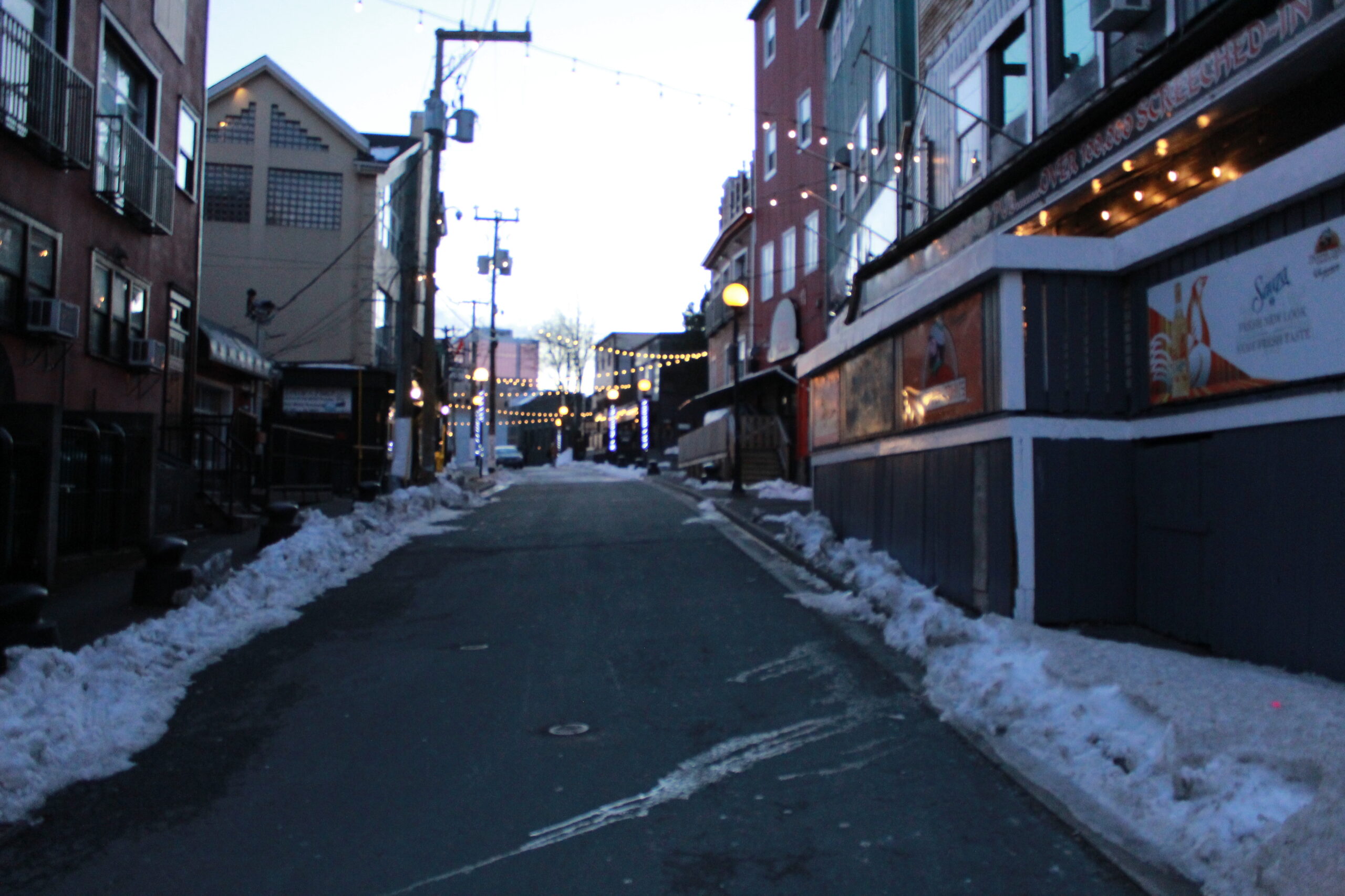 A narrow street lined with bars, clubs and breweries. Snow is accumulated on either side of the curbs. Its a clear day as the sun is setting in the sky, dusk. The road is empty expect for one person off in the distance. 