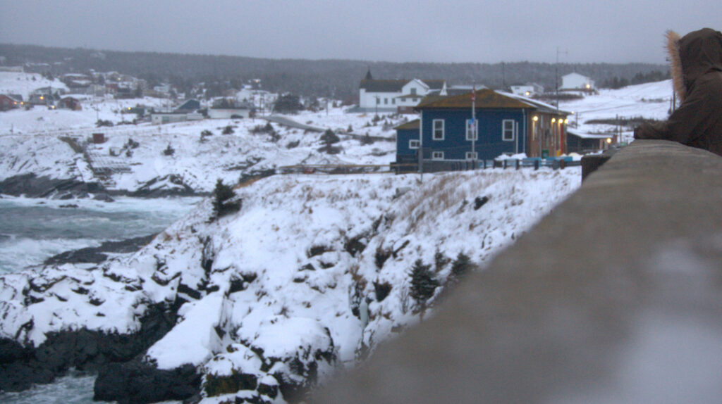 A woman is leaning over a concrete railing and looking at the rough sea below her. The Pouch Cove firehall is in the background.