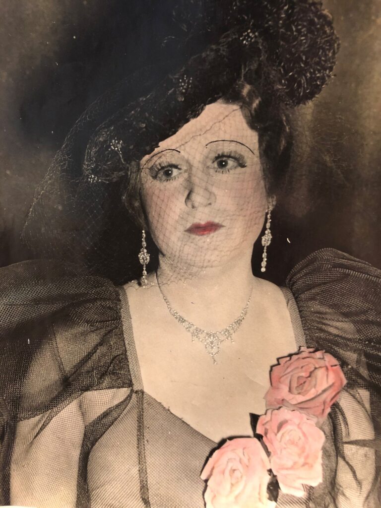 Ross Hamilton, a popular female impersonator during the world wars, is pictured in costume. He is wearing a black fascinator hat with a lace piece that falls over his face, dangling diamond earrings with a matching necklace and a black lace dress with three pink roses attached to the neckline. He is wearing red lipstick and thin, drawn on eyebrows. 