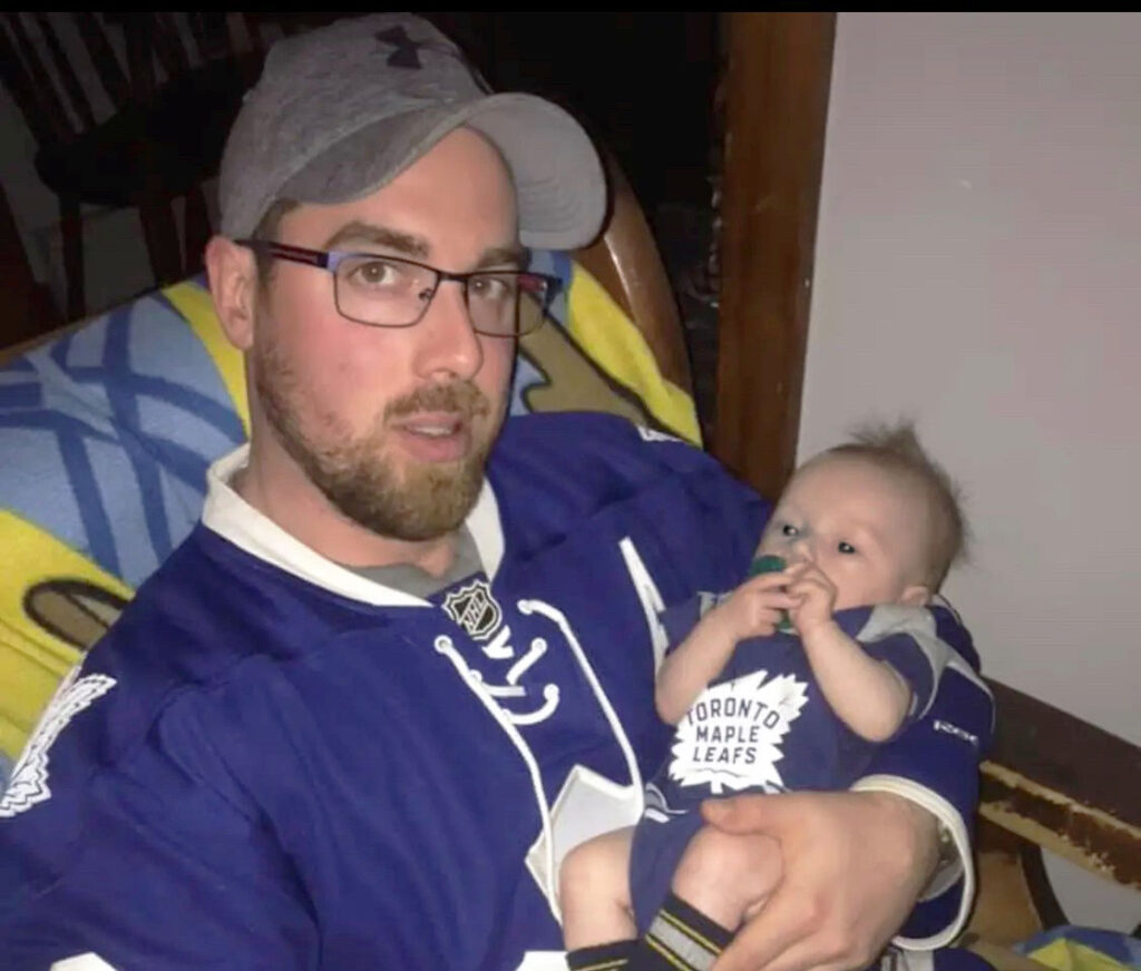 man with beard and glasses wearing a Toronto maple leaf's jersey holds a baby around four months old also wearing the same jersey.