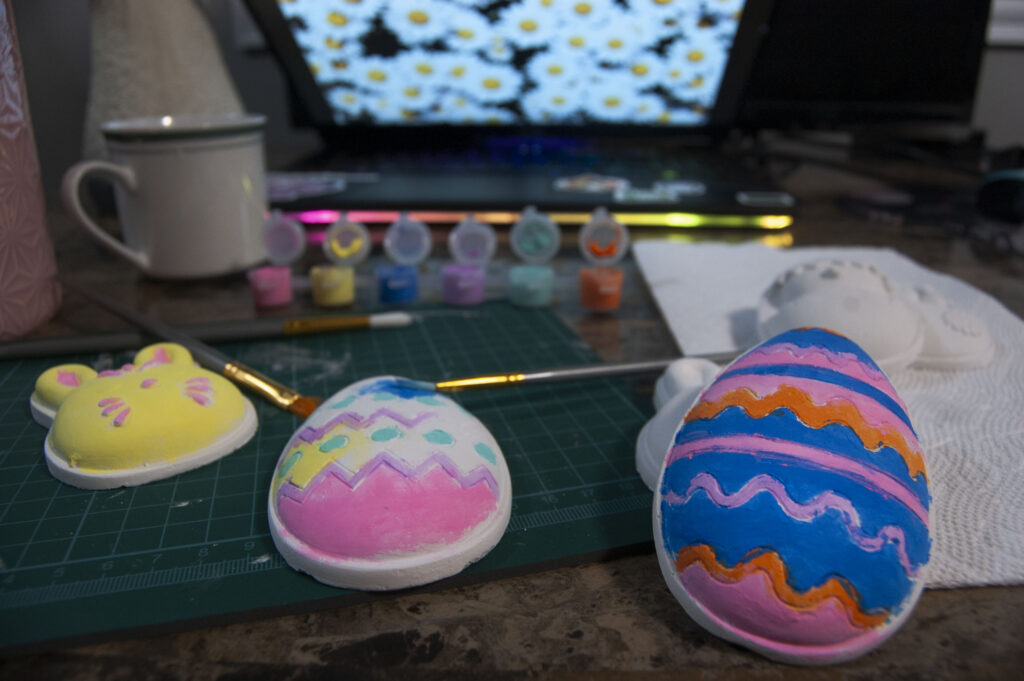 There is a pink, blue, and orange egg beside one that is half painted with pink, purple, yellow, and blue colors. Beside that one is a bunny face, painted with yellow and pink. Behind those are paintbrushes and the paint caps, which is next to a white tea cup full of water. In the background is a laptop screen with white daisies on it. 