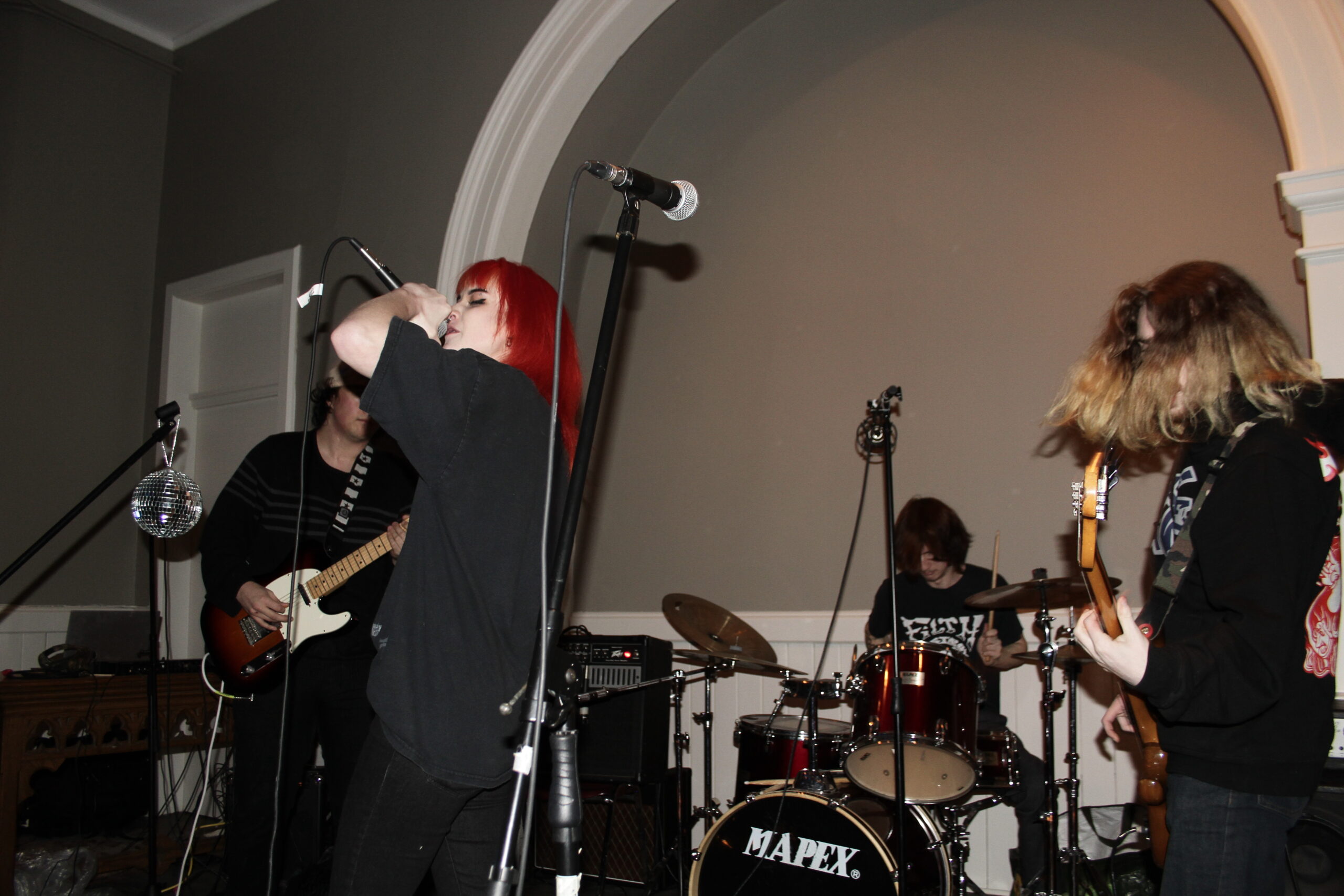 A punk rock bands performs in the Gower Street United Church. The wall is grey and there is a white arch in the middle of it. A silver microphone on a black stand is seen in the foreground. The singer yells into an upraised microphone. She has bright red hair. There are two guitarists, one of which, who is standing on the right, has his blonde hair covering his face. There is a man sitting at a drum kit behind the other three band members. 
