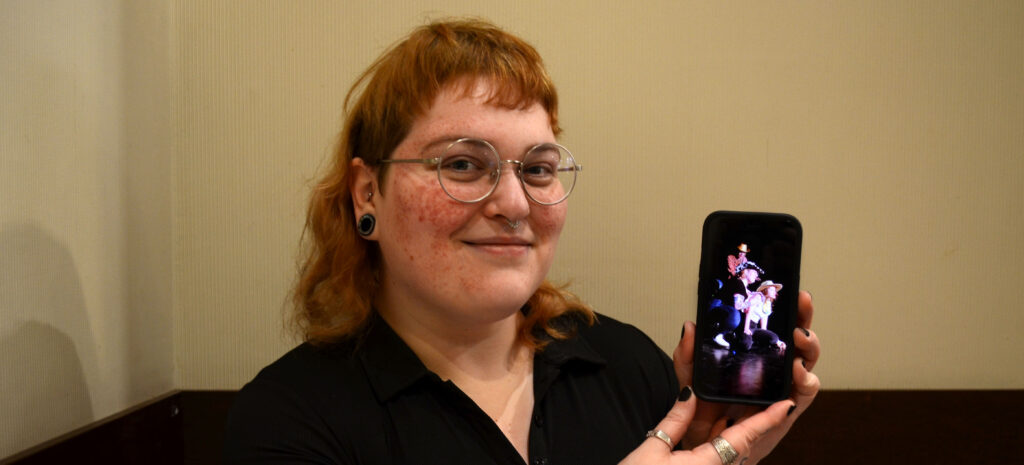 Stef Curran smiles at the camera in front of a beige wall with dark wood border at the bottom. She has a strawberry blonde mullet and round glasses. Her skin is pale with freckles. She is wearing a plain black shirt with a slight v-neck. She is holding up a picture of her comedy trio, Mom's Girls, on her iPhone. All three people in the image are wearing cowboy hats.