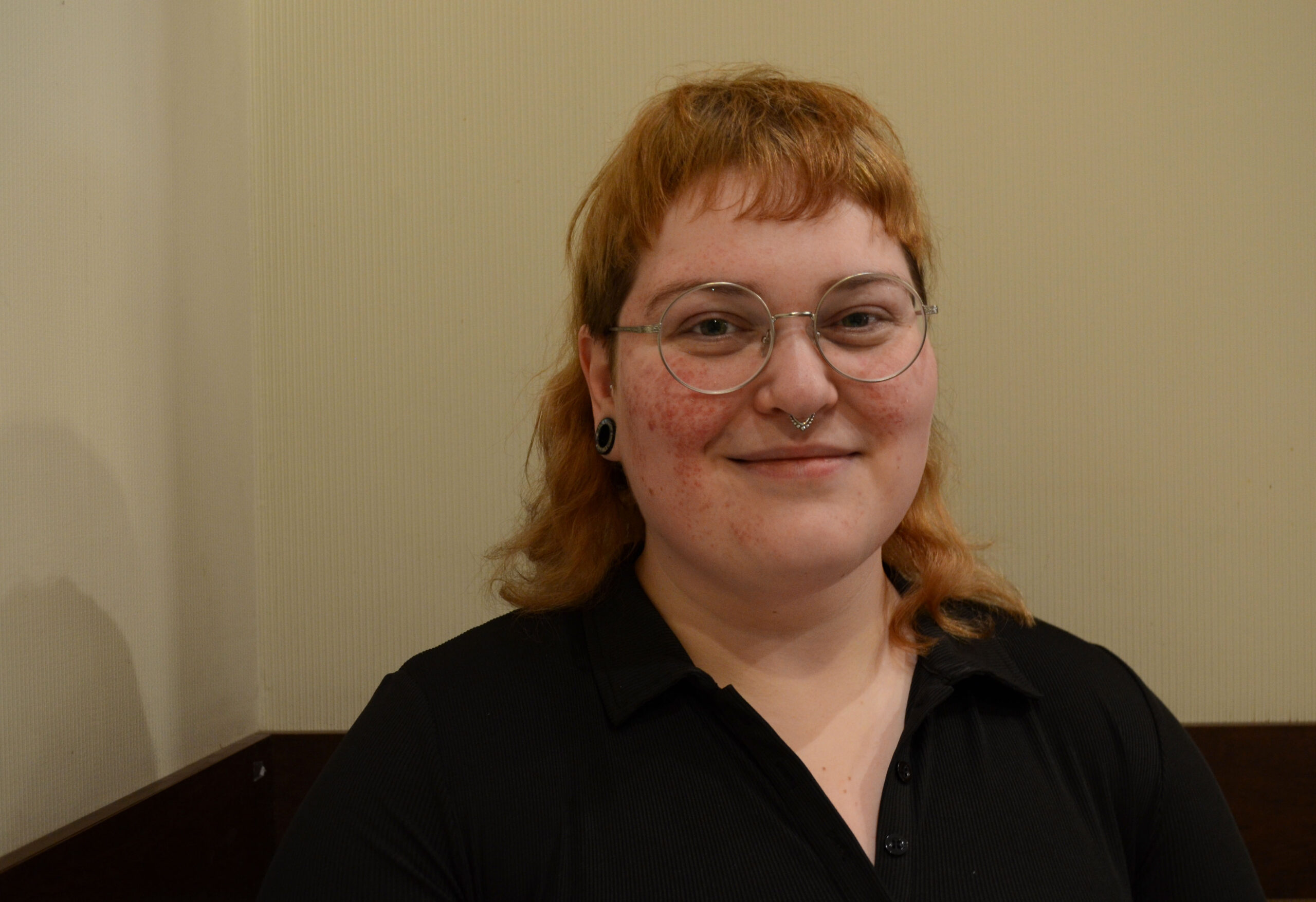 Stef Curran smiles at the camera in front of a beige wall with dark wood border at the bottom. She has a strawberry blonde mullet and round glasses. Her skin is pale with freckles. She is wearing a plain black shirt with a slight v-neck.