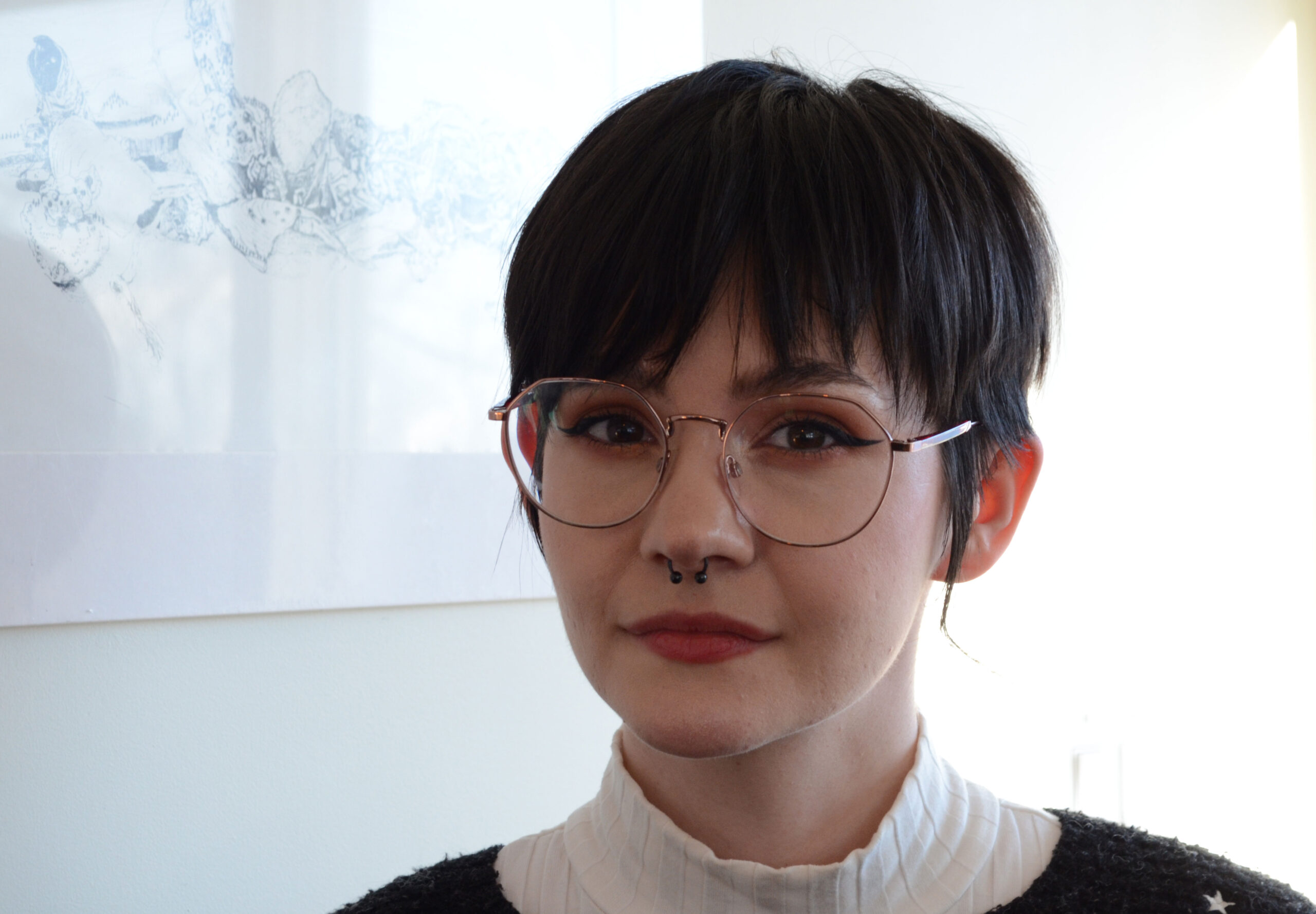 Krys Burton, a musician, is sitting inside the Battery Café. Kris has a back pixie cut, round silver-framed glasses, and a black septum ring. She is wearing pink eyeshadow with black winged eyeliner. She is caucasian. She is wearing a white turtleneck shirt under a grey sweater with white embroidered stars. This is a close-up shot, so we can only see the white wall and a blurred-out drawing behind her. 
