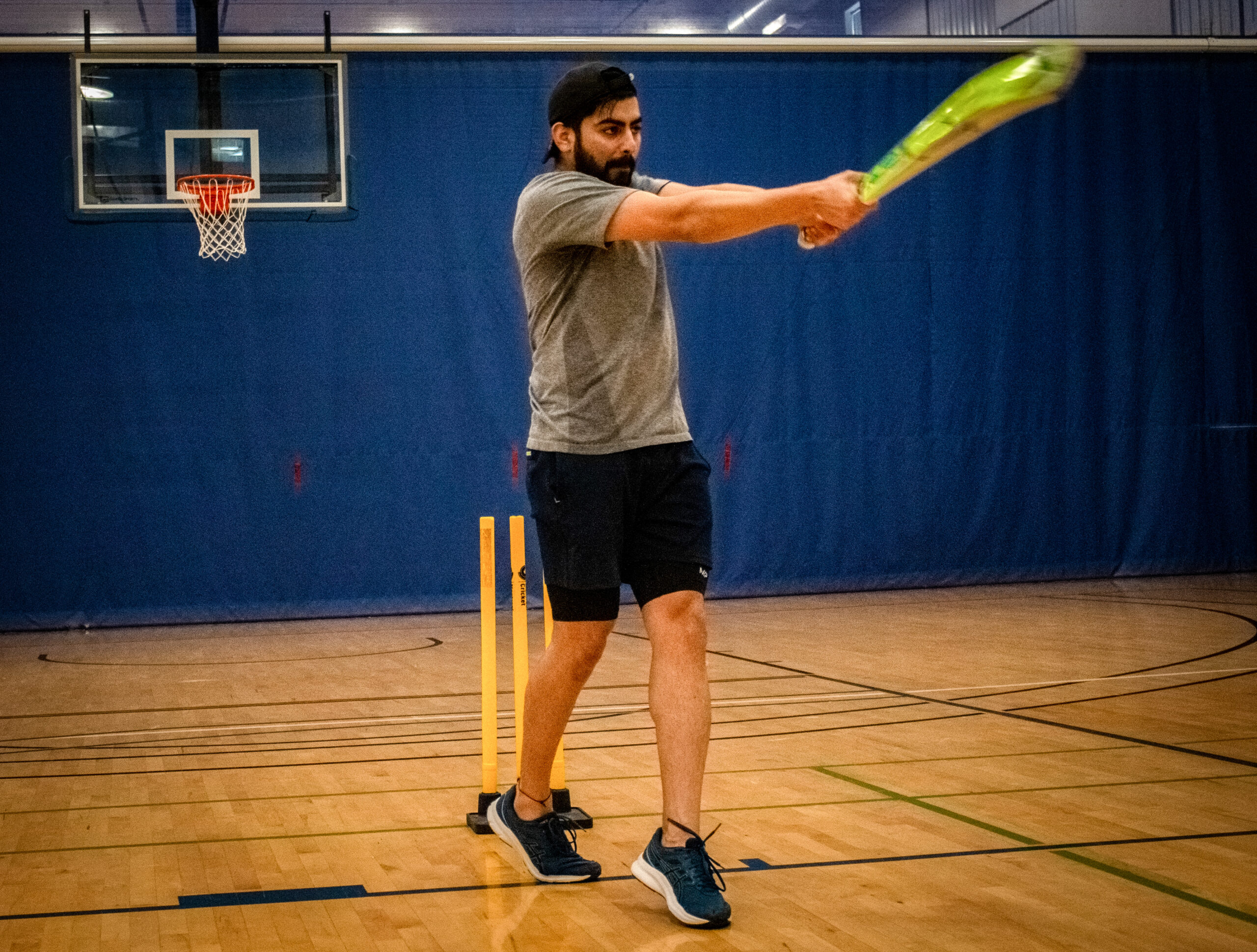 A man swings a cricket bat in a gymnasium with a basketball net in the background.