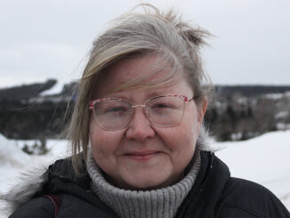 Blond woman with glasses standing infont of hills of snow. In the background there are trees,