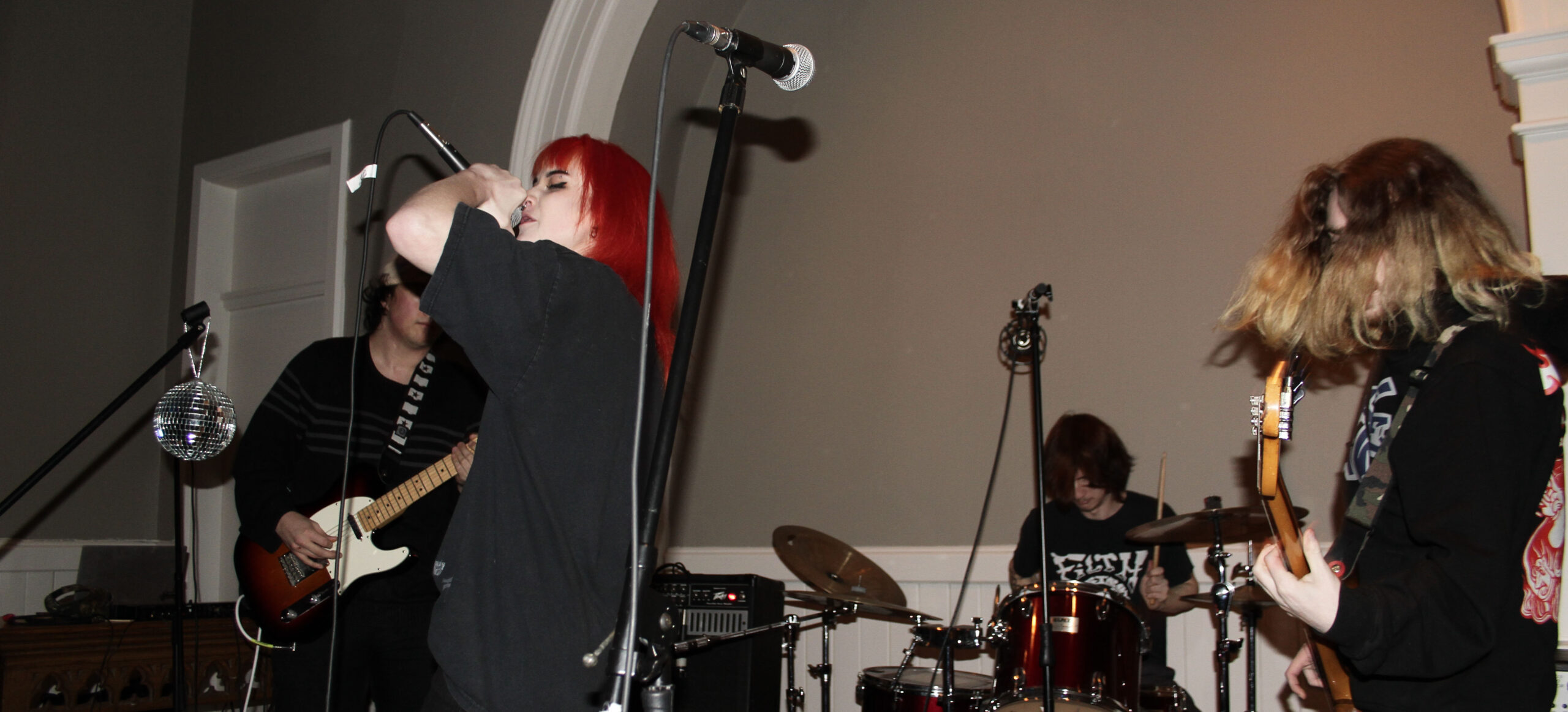 A punk rock bands performs in the Gower Street United Church. The wall is grey and there is a white arch in the middle of it. A silver microphone on a black stand is seen in the foreground. The singer yells into an upraised microphone. She has bright red hair. There are two guitarists, one of which, who is standing on the right, has his blonde hair covering his face. There is a man sitting at a drum kit behind the other three band members.