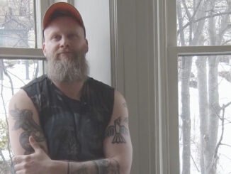 A man with a beard and many tattoos stands against a wall in the Anna Templeton Centre with his arms crossed. He is wearing an orange ball cap and a black sleeveless shirt that he designed himself.