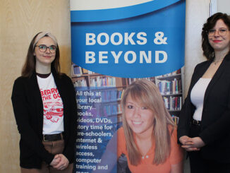 Emma Craig(left) and Sarah Bartlett (right), both wearing black, stand in front of a blue banner that says "Books and Beyond"