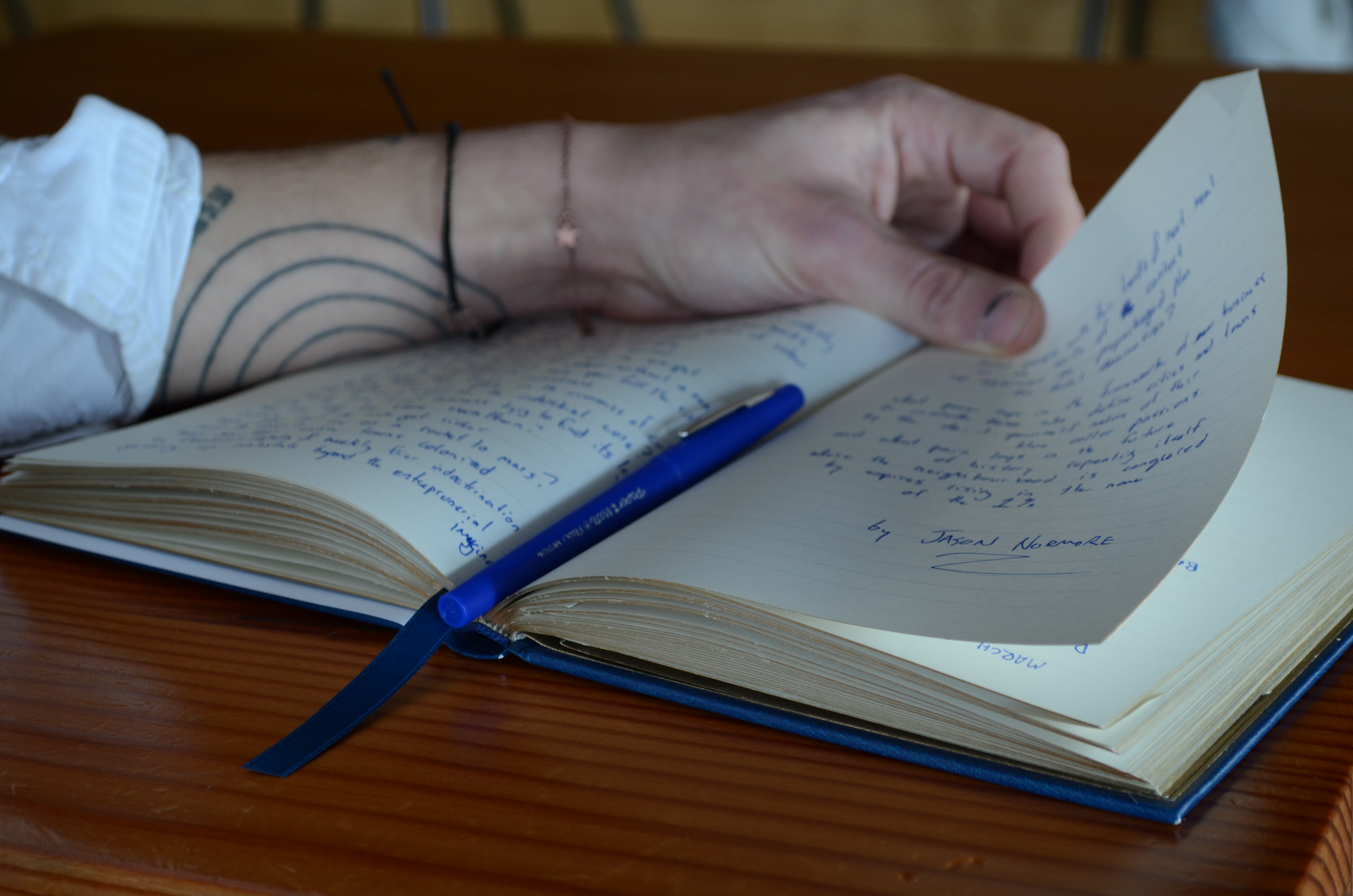 Jason Normore flips through his poetry notebook. The pages are marked in blue ink, and a blue pen lays in the centre of the book. Tattooed lines on his arm can be seen from this angle. 