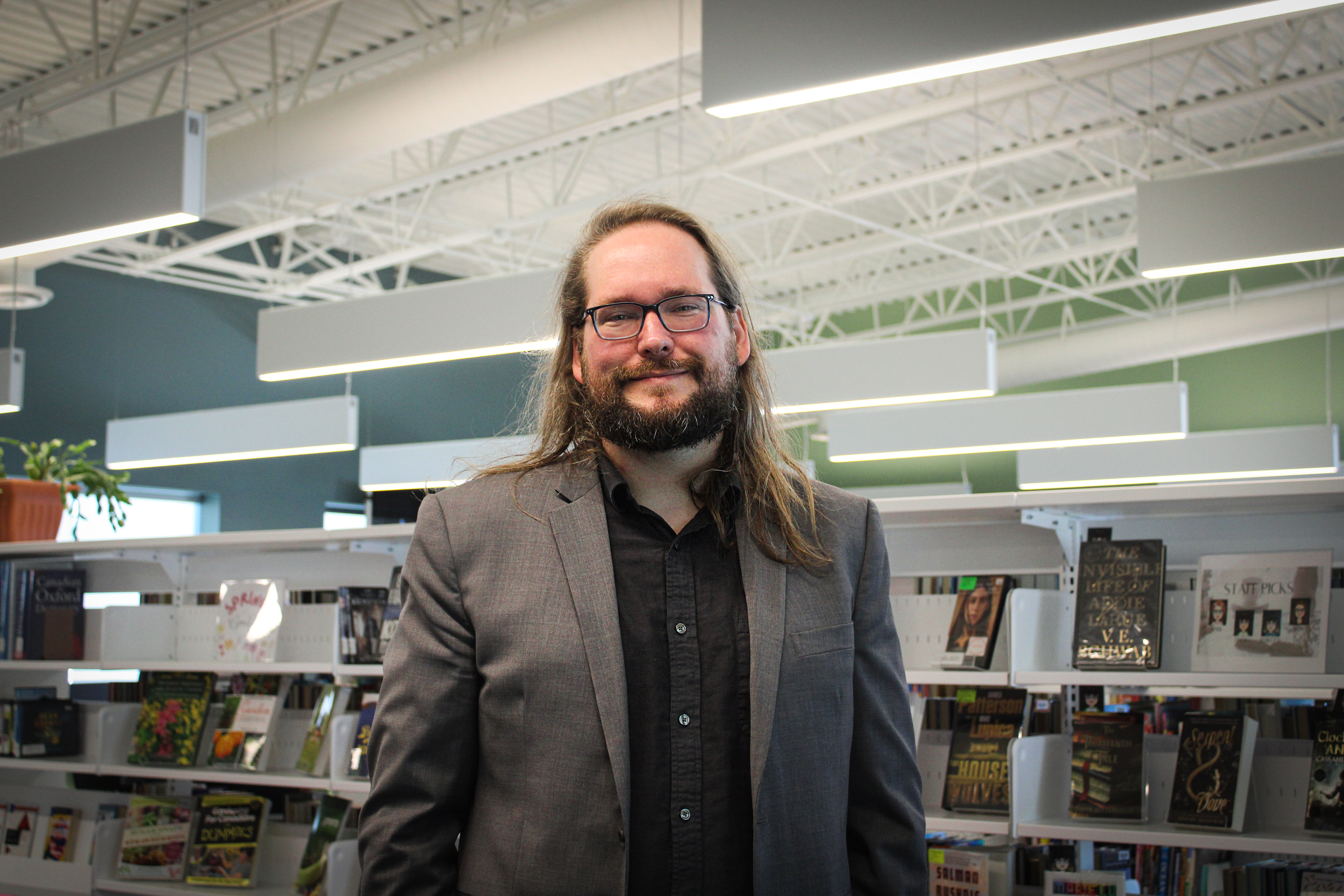 Andrew Lockhart, in black shirt and grey suit stands in front of CBS library shelves. He has shelves with books in the background and ceiling lights to his top.