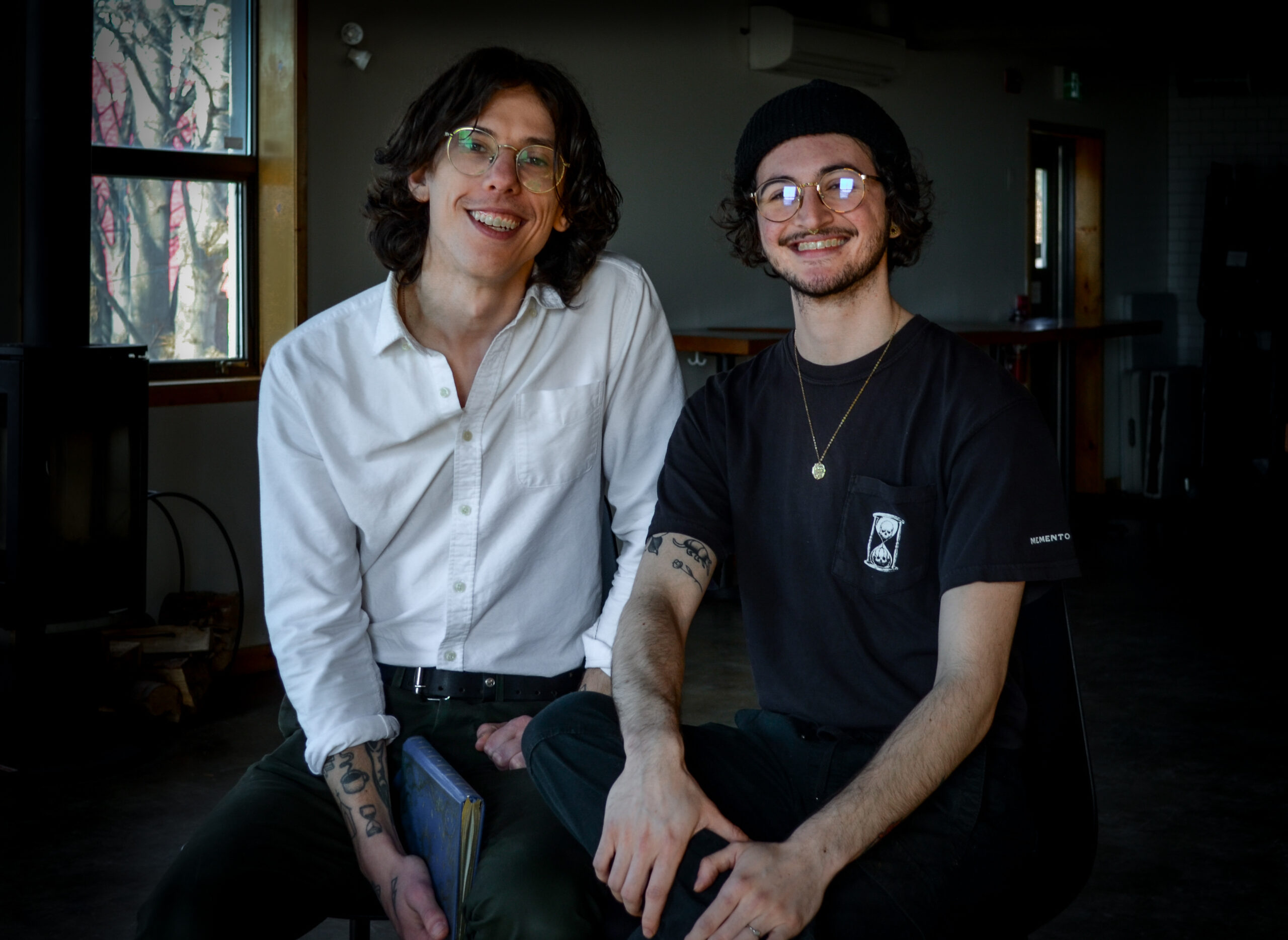 Jason Normore and Lincoln Ripley sit pn stolls at the second floor of the Bannerman brewery. Jason is wearing a white button-down collared shirt with grey pants. He has several arm tattoos, messy brown hair, and round glasses. Lincoln is wearing a black beanie hat, a dark grey t-shirt with an hourglass on the chest, and round glasses. A window is seen in the background.