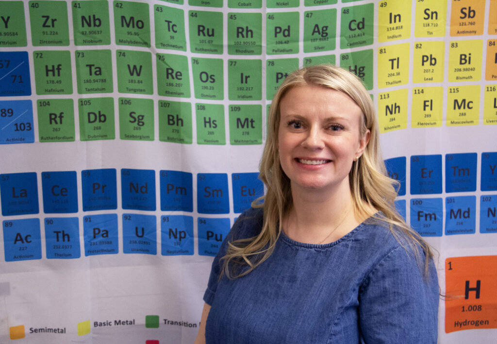Heidi Kavanagh stands in front of a colorful periodic table. She encourages deep learning through hands-on work.