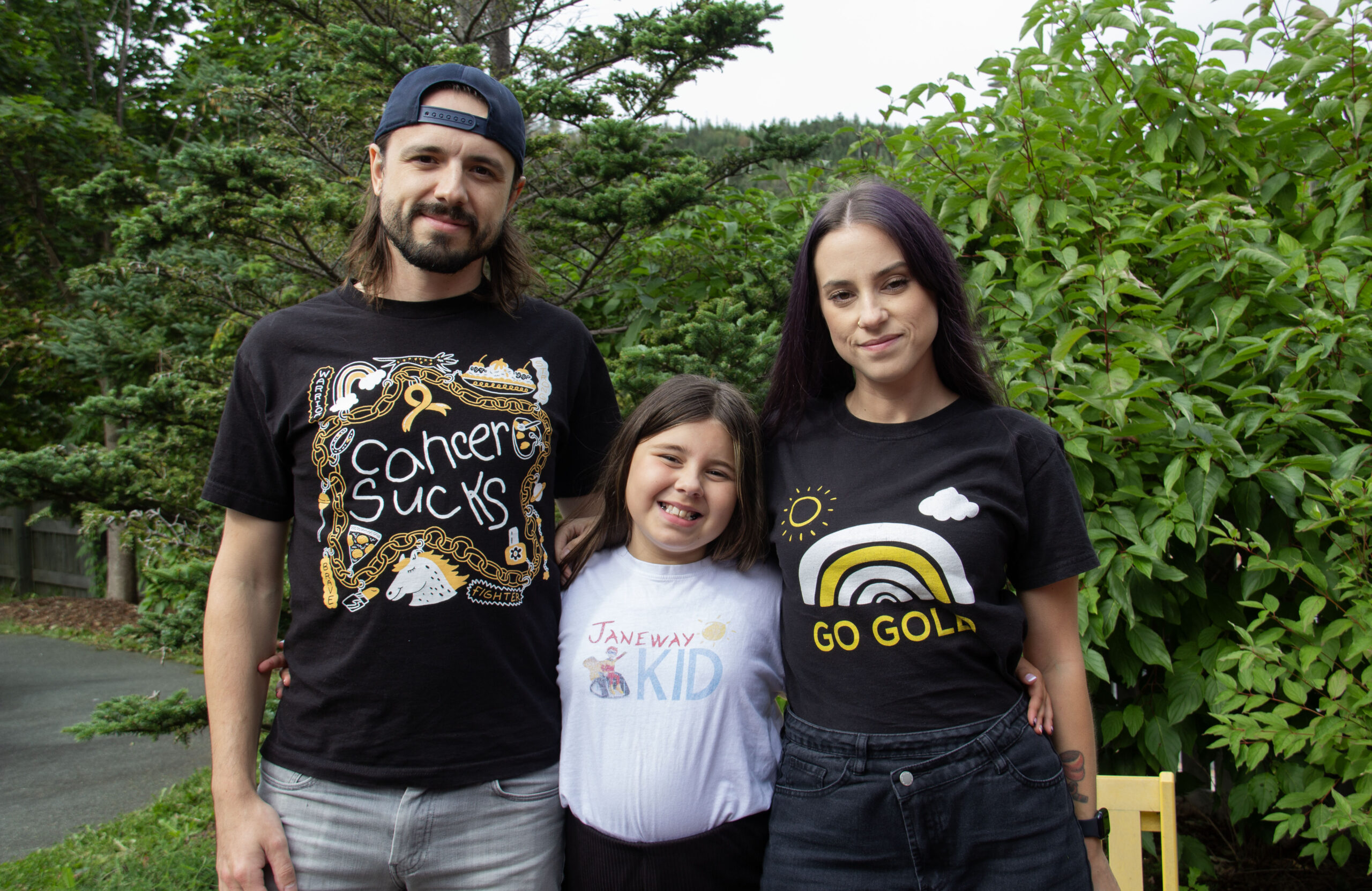 Abigale Latoszeck stands wearing a white shirt that says "Janeway Kid". On her left is her dad, wearing a black t-shirt that says "cancer sucks", and on her right, her mom is wearing a black t-shirt that says "go gold". They are outside with a background of green trees