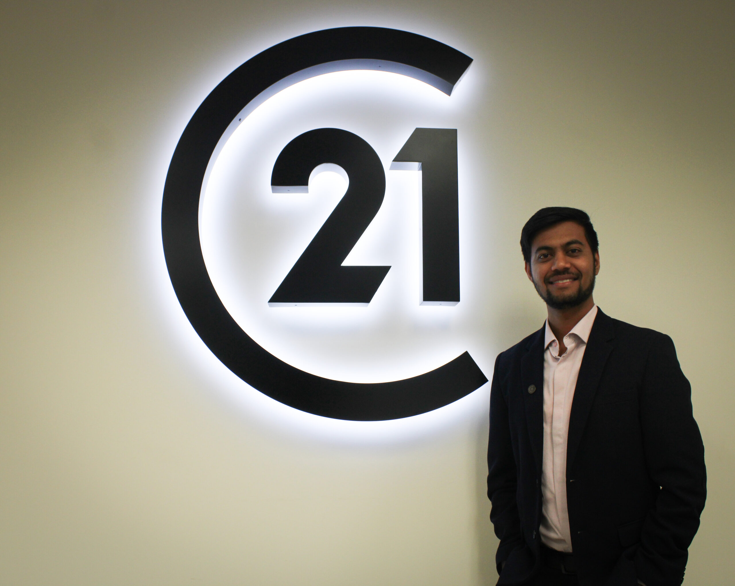 A real estate agent stands in front of Century 21 logo on the wall. He is wearing a black suit and smiling at the camera.