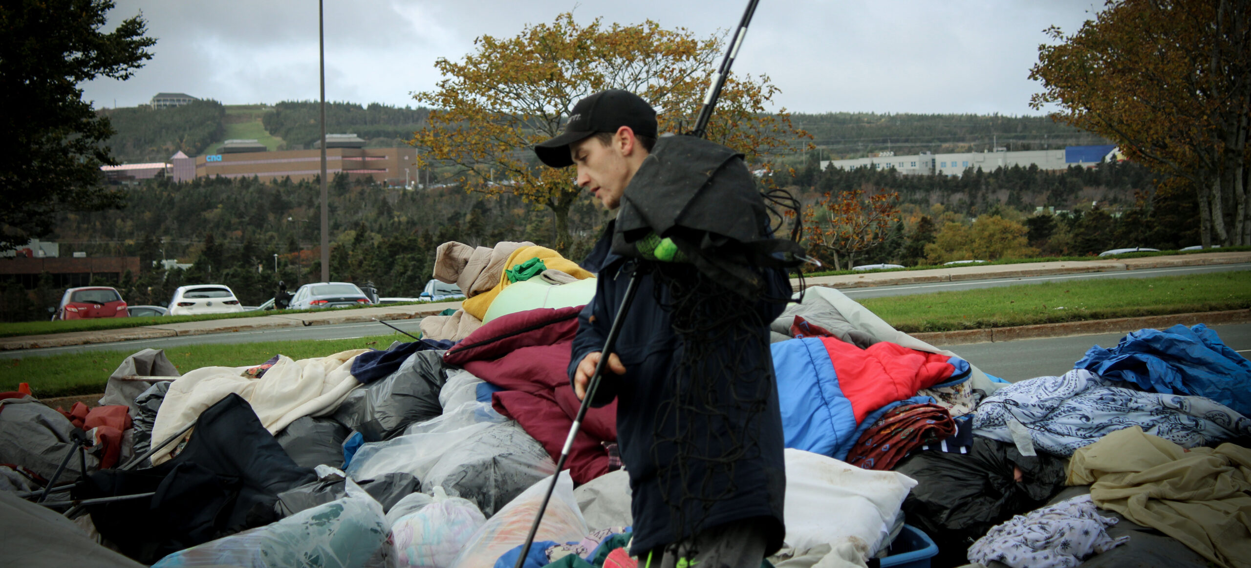 A tent city resident, in a black jacket searches for personal items from a huge pile of debris. The pile has many blankets, clothes as well as tent equipment.