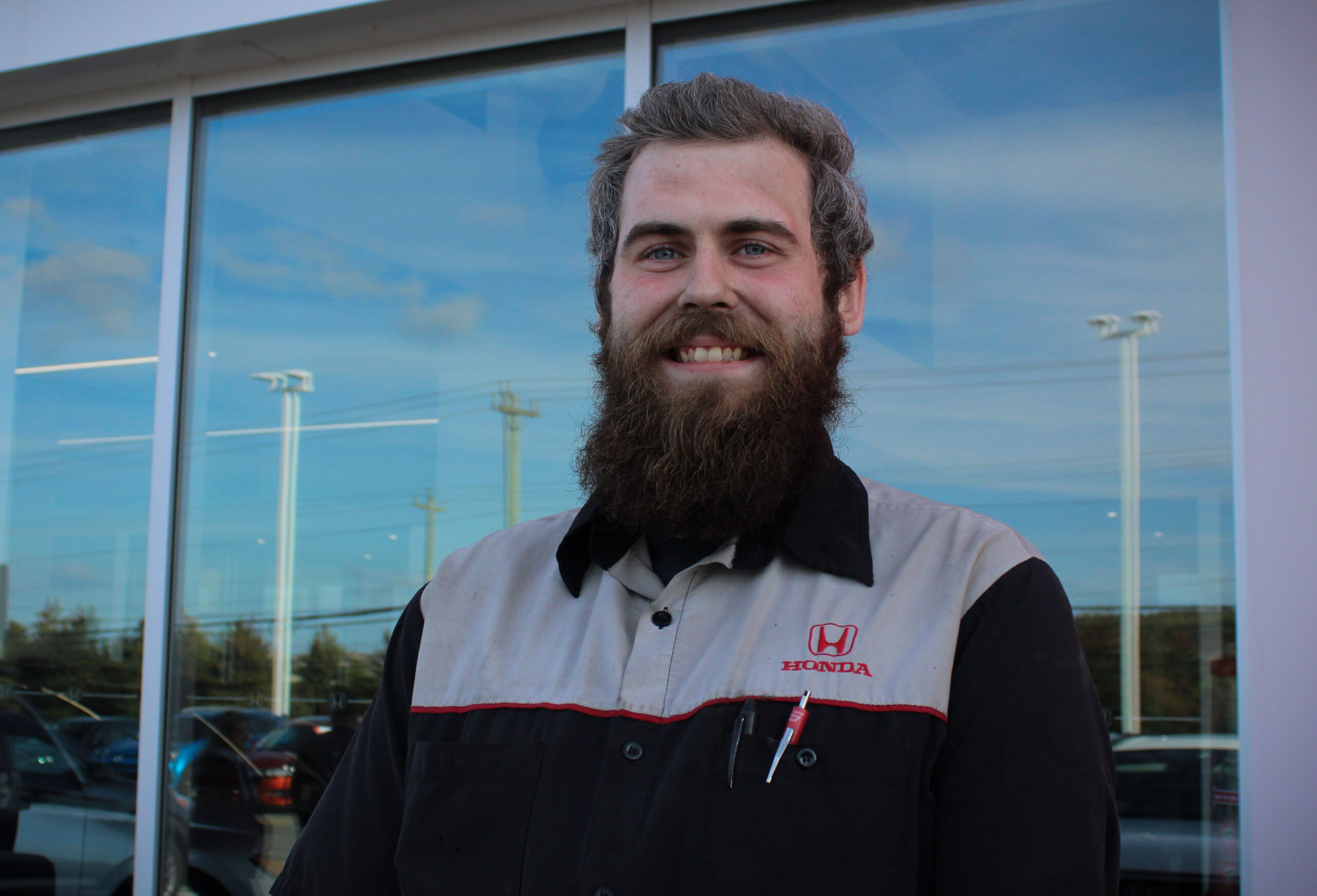 A mechanic in black and grey Steele Honda uniform stands infant of a glass building smiling at the camera. He said a long beard as well.