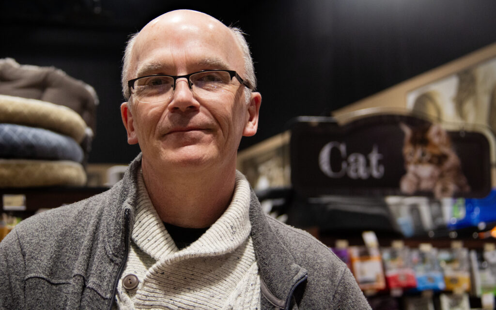 A man smiles at the camera, with the "cat" aisle of a pet store in the background