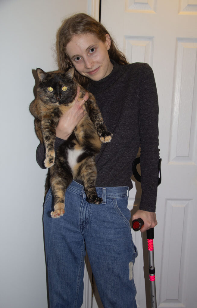 A woman stands using a crutch with her left hand, holding a tortoiseshell cat in her right.