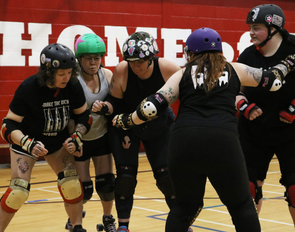 A girl with red hair and purple helmet braces with three other people to stop a girl with a green helmet cover breaking through the group.