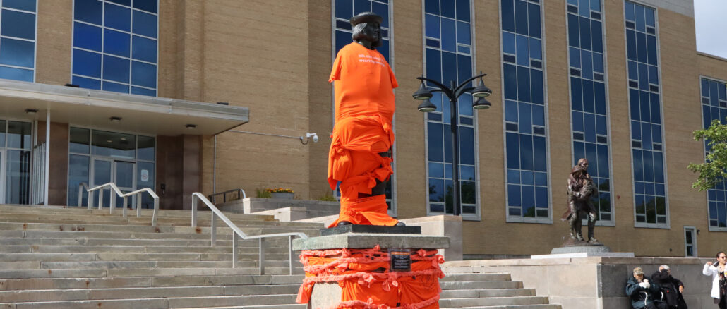 image shows John Cabot's statue in front of the confederation building wrapped in orange shirts and ties.