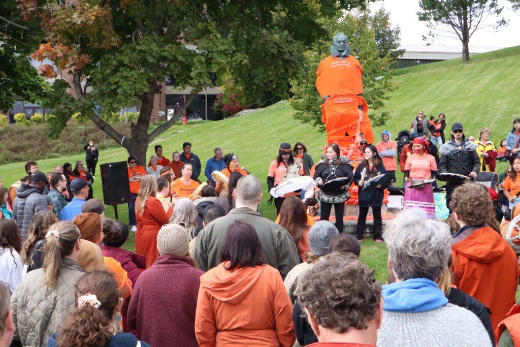 Image shows people gathered around a statue wrapped in orange as they sing and drum. 