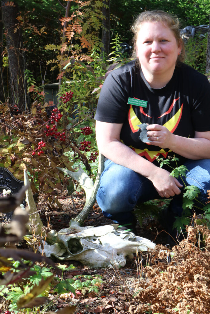 A blonde woman poses outdoors holding a bat and is next to a moose skull