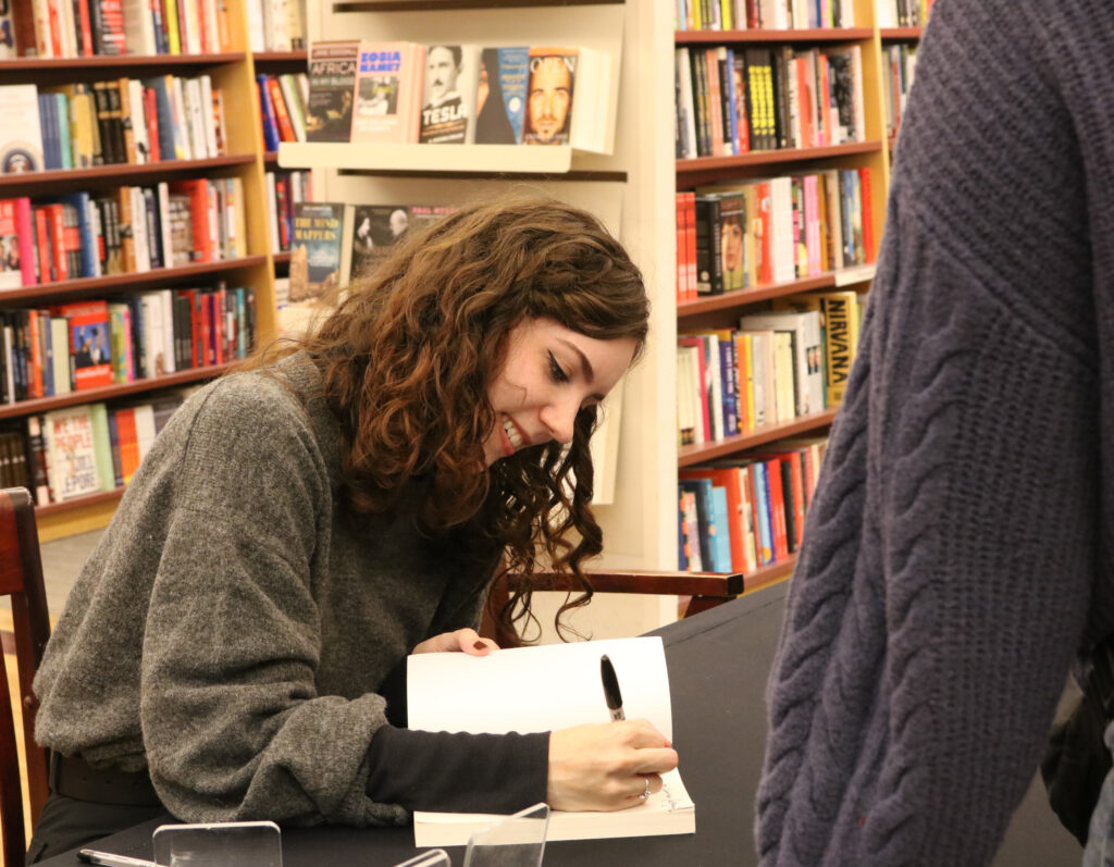 a girl with curly brown hair named Mackenzie Nolan leans over her book with a smile and signs it. 