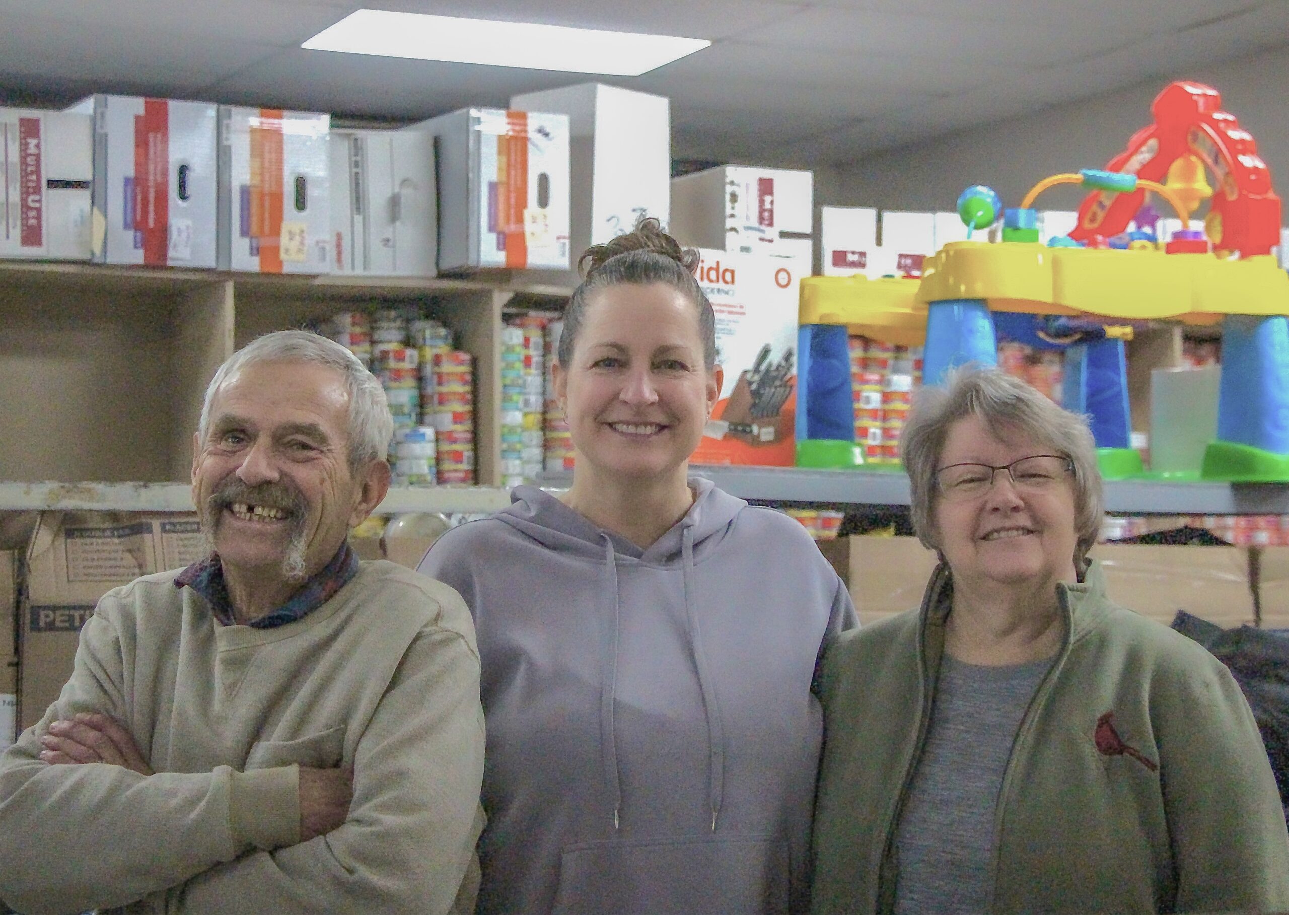 Three members of the CBS-Paradise Food Bank are smiling ear to ear. The three stand in front of boxes, canned goods and toys.