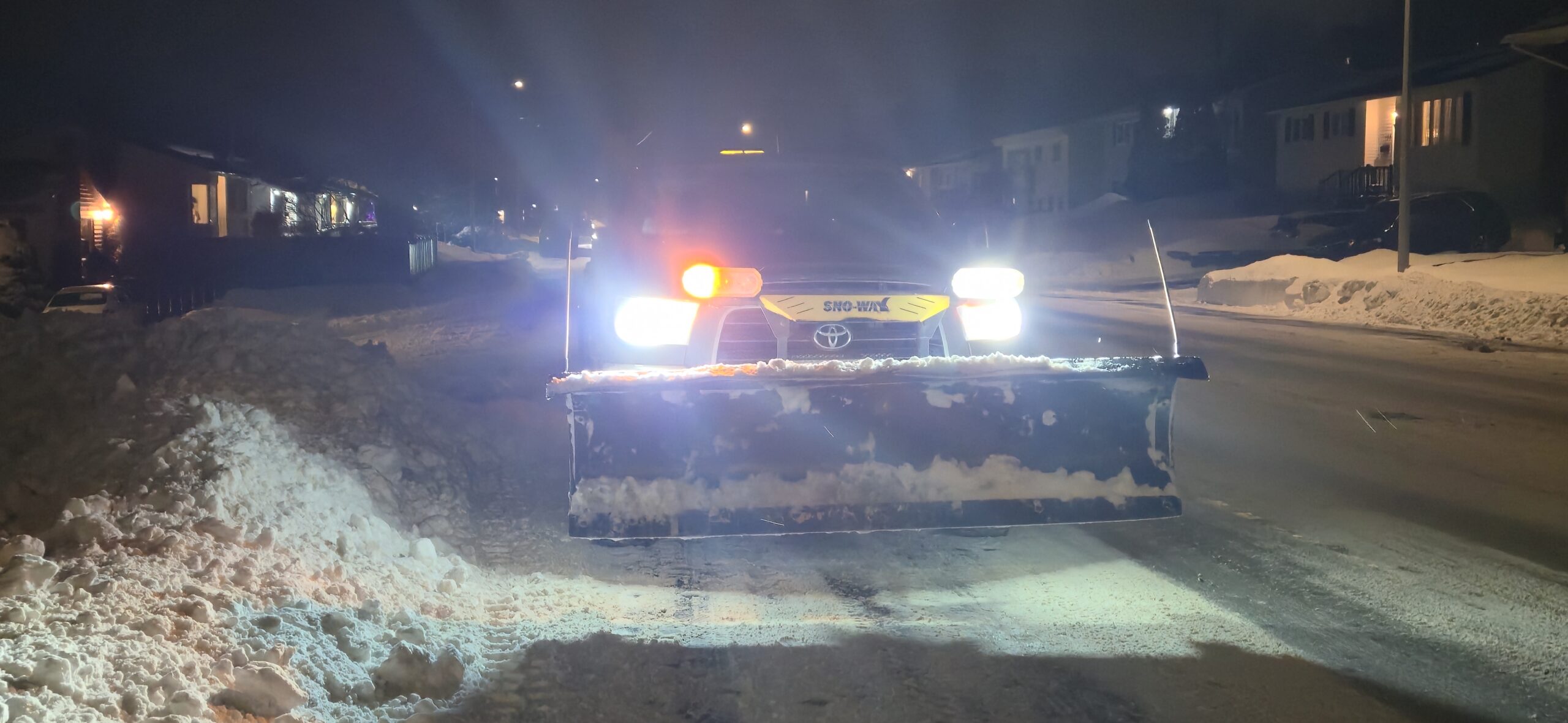 A plow clears snow on a dark street in St. John's, headlights glaring into view. Operator Scott Ward is part of a team that clears snow for people affected by cancer.