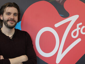 Robert Shawn, OZFM host, standing and smiling against an OZFM poster