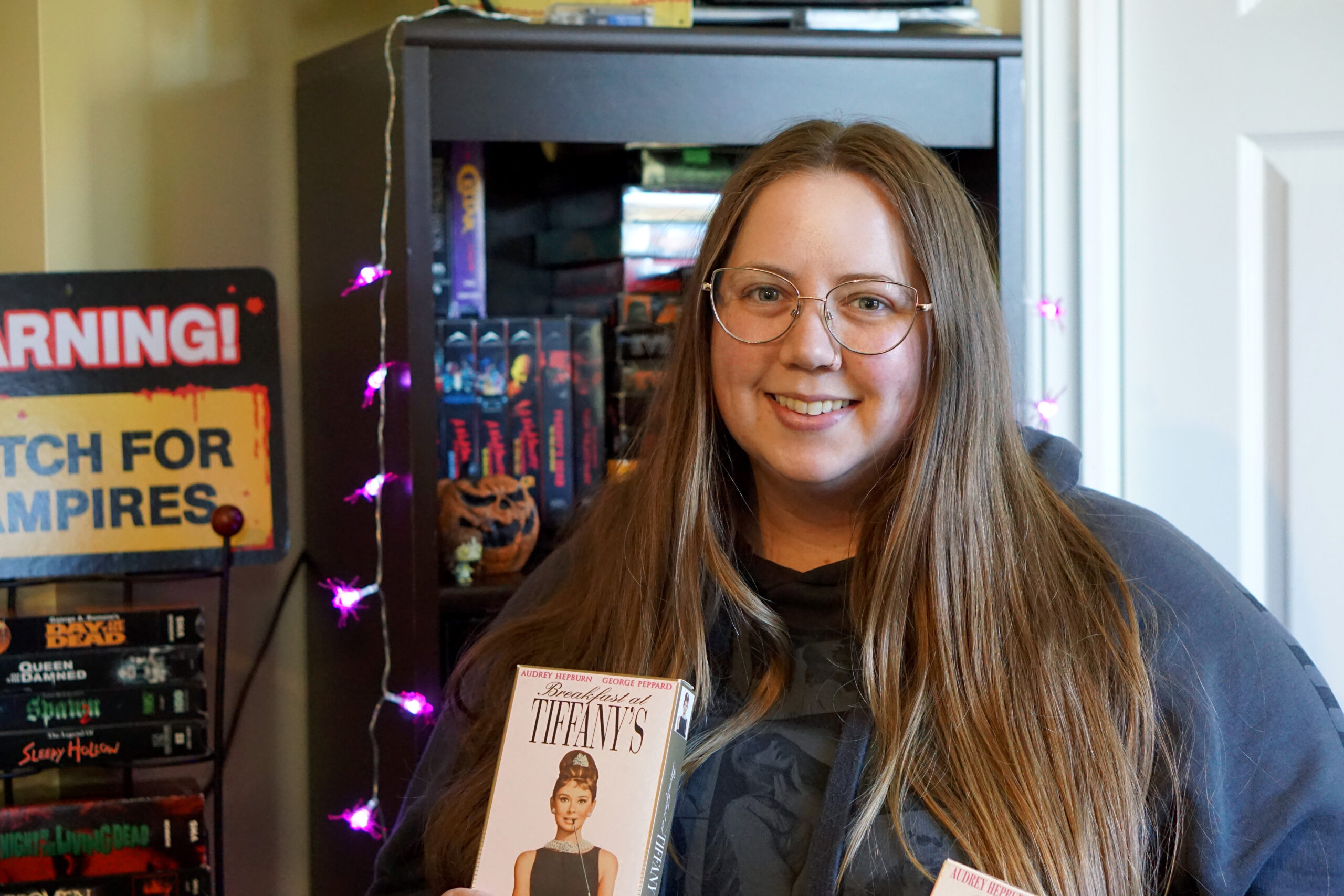 Return of the vhs tape. A woman holds a pair of VHS tapes in front of a shelf full of even more.